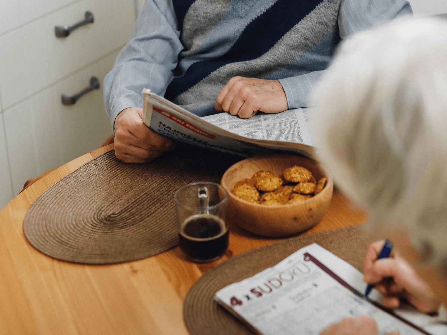 An older couple sitting at a table engaging their minds. One reads the newspaper while another completes a sudoku puzzle.