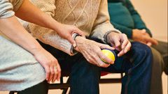 An elderly man holds a ball for physical therapy. A caring hand holds his arm.