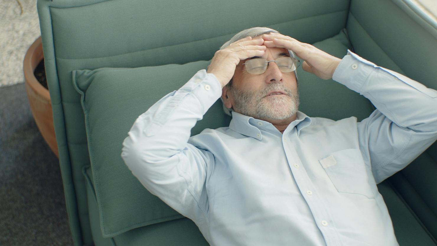 An older man lays on a couch holding his head as if dizzy or having fallen.